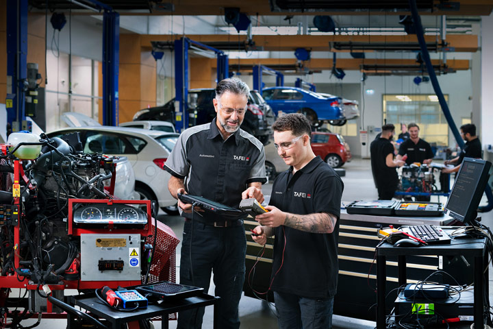 Automotive student and teacher using diagnostic tools in a workshop with cars on lifts and other students working in the background.