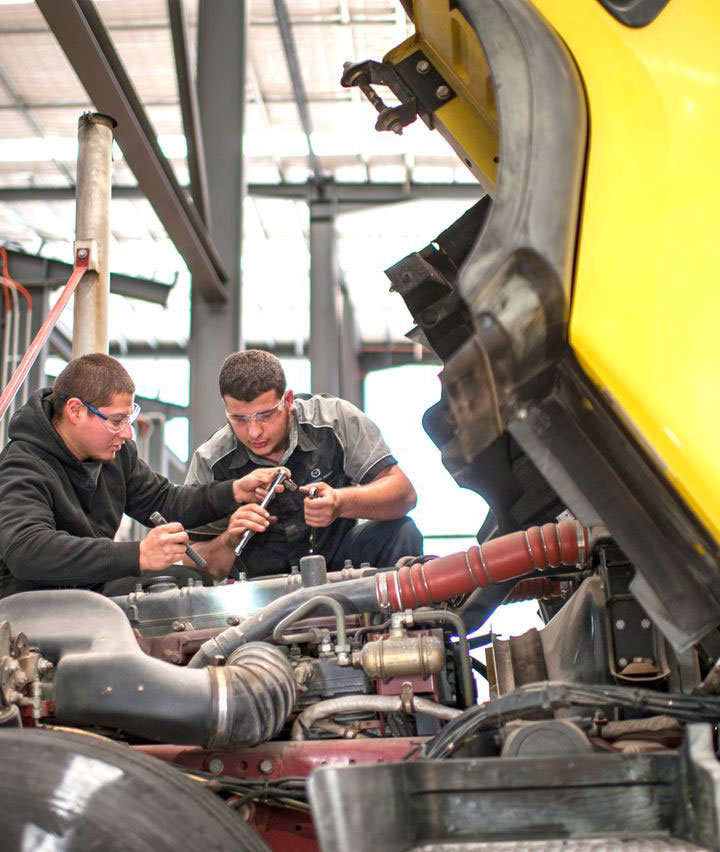 Students wearing protective eyewear work as a team on a large yellow vehicle in a purpose-built automotive trades and services space. The image showcases a practical learning environment.