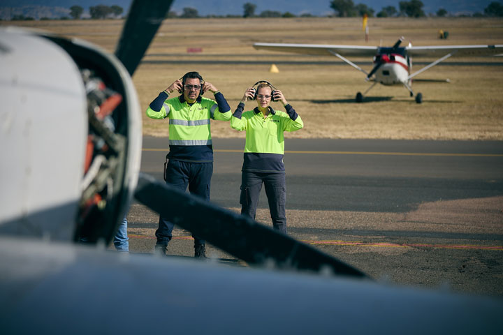 Two aviation students in high-visibility shirts stand on an airfield, adjusting their headsets as they prepare for aircraft operations. A small airplane is visible in the background.