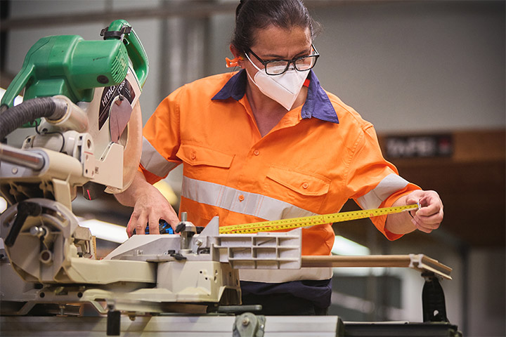 Flooring technology student measures a piece of timber using a tape measure while operating a drop saw in a workshop, wearing an orange high‑visibility shirt and safety 