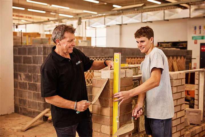Bricklaying student and teacher work together in a training workshop, using a spirit level and trowels to check alignment and lay bricks on a partially built wall.