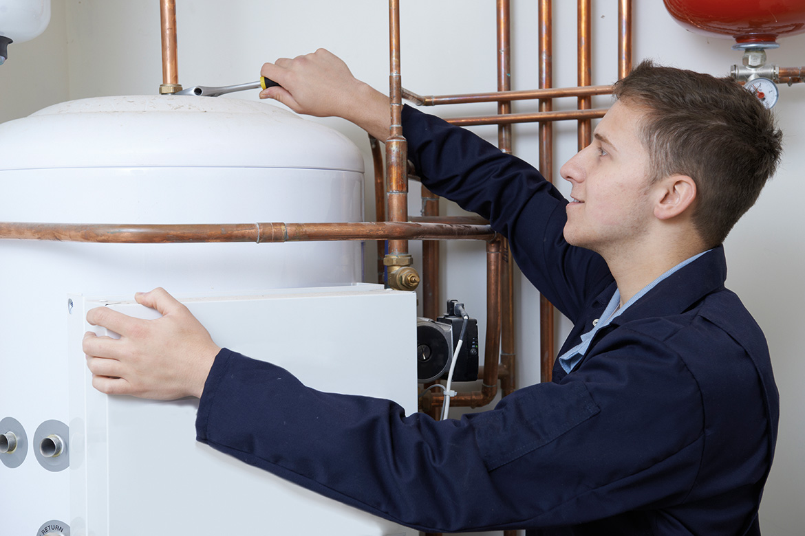 A young technician in a blue coverall works on a boiler system, adjusting a component while examining the pipes and tank.