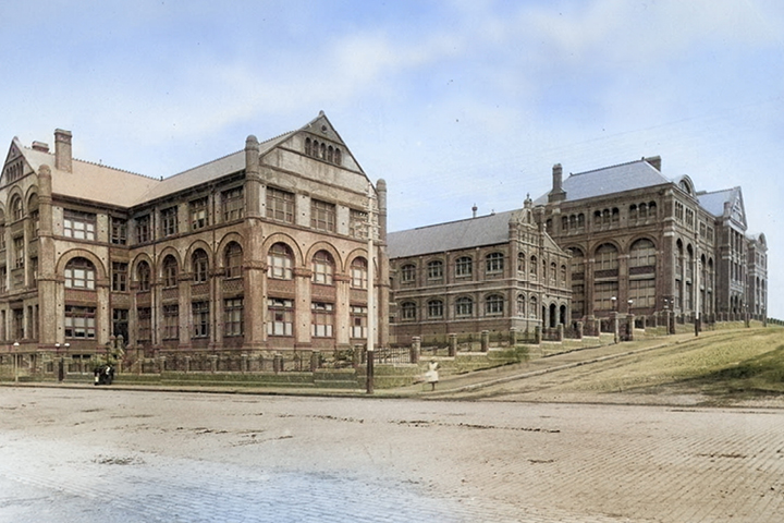 Students stand on a cobblestone path beside brick and sandstone historical buildings. A neat hedge borders the area.