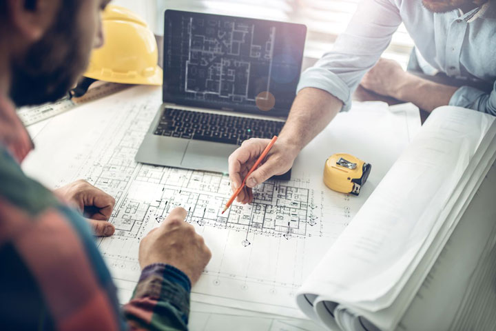 An overhead shot depicts two men collaborating on building plans, as one makes notes. They review blueprints next to a laptop showing another design. A hard hat and measuring tape lie nearby.