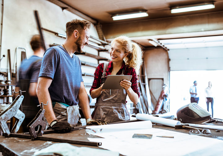 Two young workers in overalls collaborate in a workshop. A woman holds a tablet, and a man stands by a workbench with tools and blueprints. Teachers observe in the background.