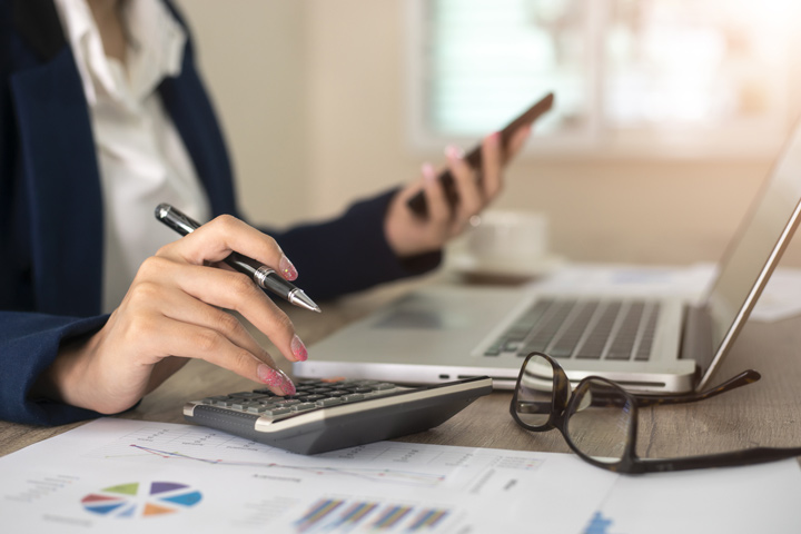 The image shows a person in a professional setting using a calculator while holding a smartphone. Their desk is filled with a laptop, financial charts, and papers, suggesting they are working on financial calculations or business reports. The person is wearing a dark jacket, and a pair of glasses rests nearby, adding to the sense of focused, detailed work.