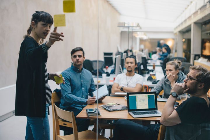 A woman leads a discussion, pointing at sticky notes on a glass wall, while four colleagues sit around a table, listening attentively. Laptops and notebooks suggest a collaborative work environment.