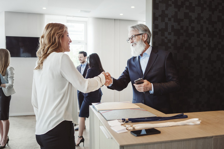 Two individuals shaking hands across a counter during a business meeting, with others conversing in the background in a modern office space featuring large windows, light-colored walls, and a flat-screen TV.