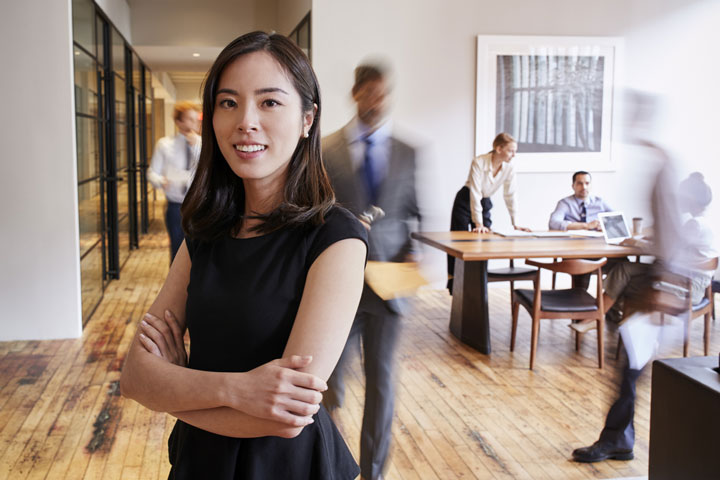 A confident young woman stands with her arms crossed in a modern office setting, while a team works in the background, creating a dynamic and professional atmosphere.