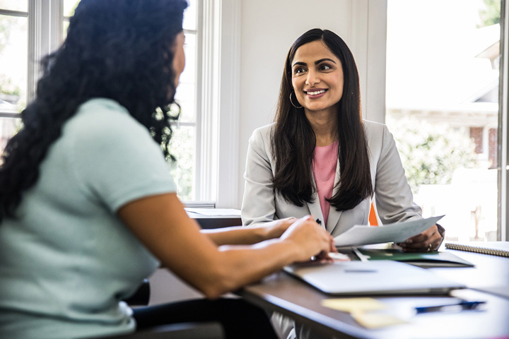 Two women talk at a table with a laptop and documents. One woman smiles and holds a paper.