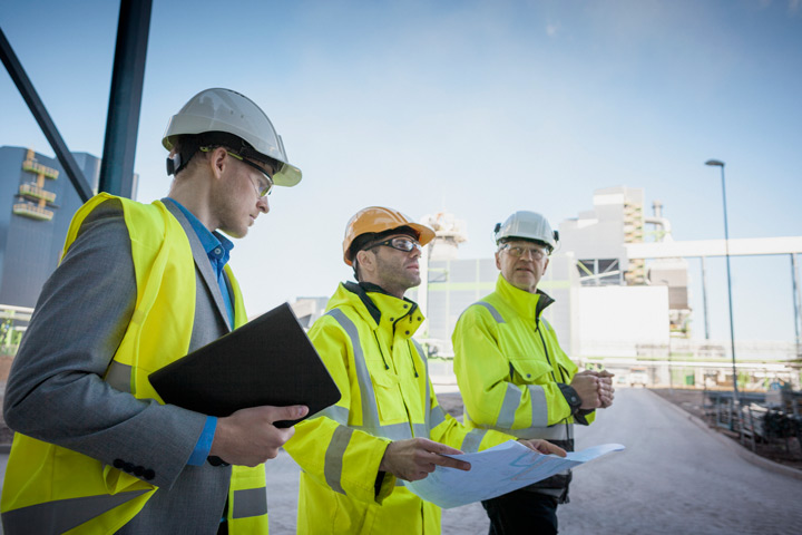 Three individuals in safety helmets and high-visibility jackets at a construction site, reviewing blueprints and discussing plans.