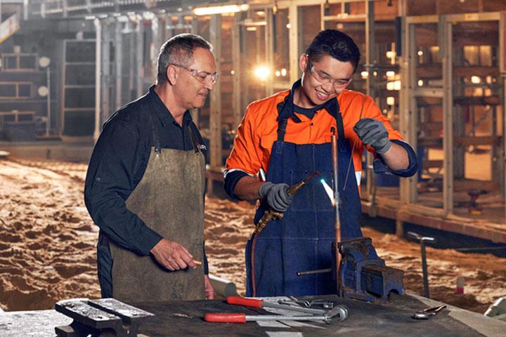 A trainer wearing safety gear supervises a student gas welding copper pipe in a workshop.