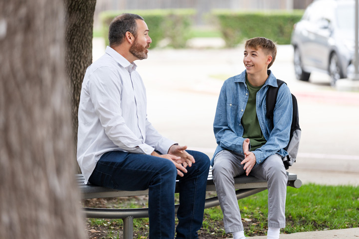 Two individuals seated on a bench outdoors, engaged in conversation in a grassy area with trees and parked cars in the background. One wears a white shirt and jeans, the other a denim jacket with a backpack.