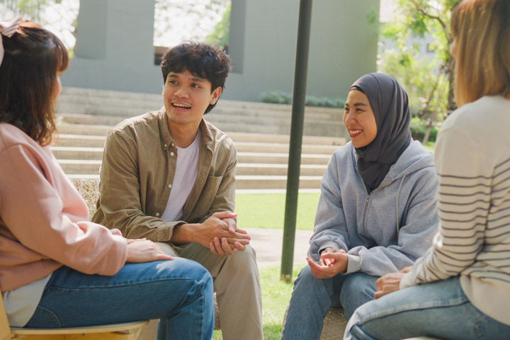A group of people are sitting in a circle have a discussion outdoors. They are smiling and engaged in conversation.