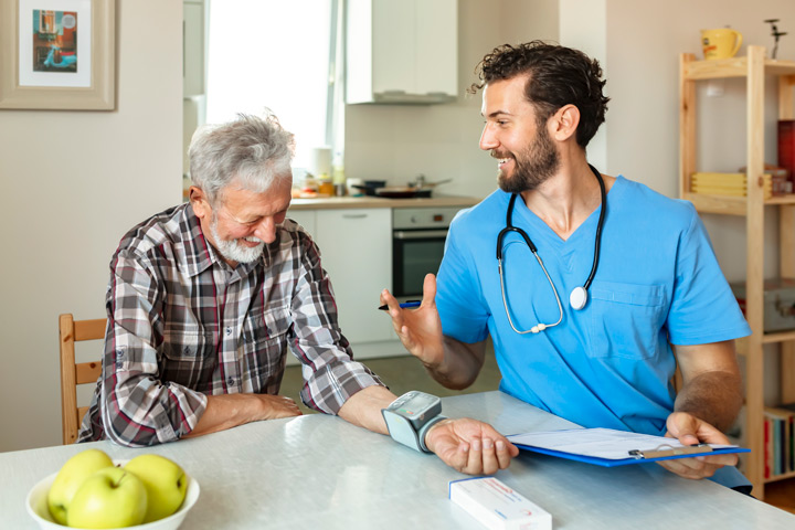 A nurse in blue scrubs with a stethoscope is seated at a table with an elderly man wearing a plaid shirt. The man has a blood pressure cuff on his arm, and the nurse is holding a clipboard and pen. Apples and kitchen appliances are visible in the background.