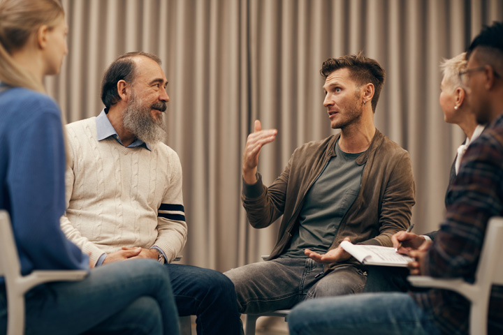 Group of individuals seated in a circle during a counselling session, with one person speaking and another taking notes, set against a backdrop of vertical curtains.