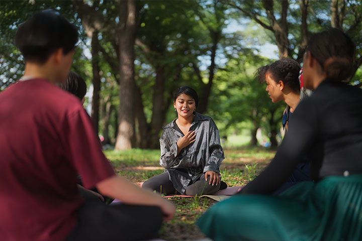 A group of people sitting on the ground in a park, all with their legs crossed. All members of the circle are looking towards one women, whom is running the circles discussion.