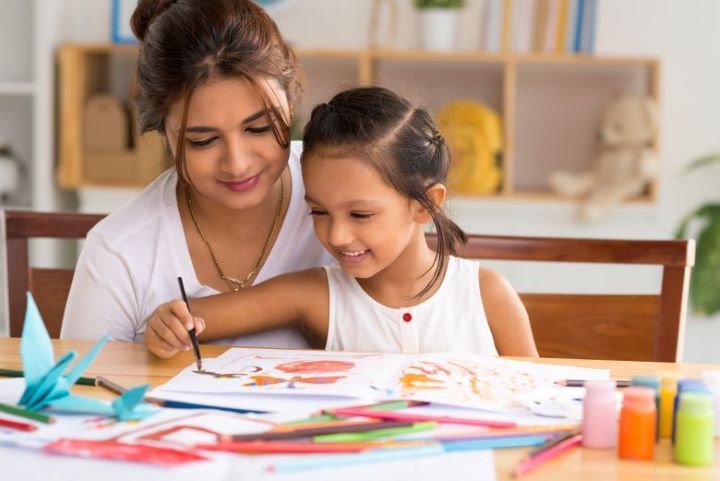A teacher and young girl sit at a table filled with art supplies, the girl holding a paintbrush.
