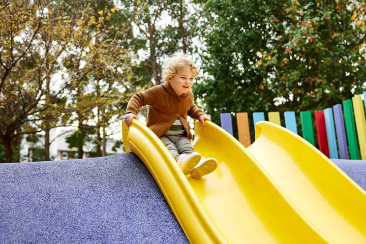 A toddler confidently perched on a slippery dip, smiling and preparing to glide down.