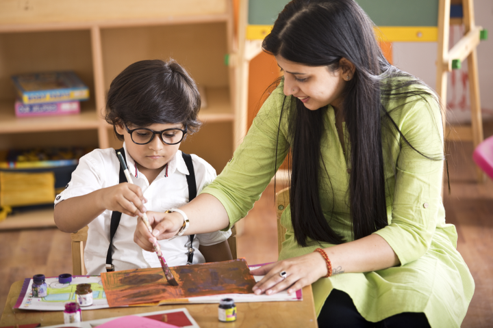 A young child is painting with the guidance of a teacher in a classroom setting. The child is focused on the artwork while the teacher helps him to hold the paintbrush.