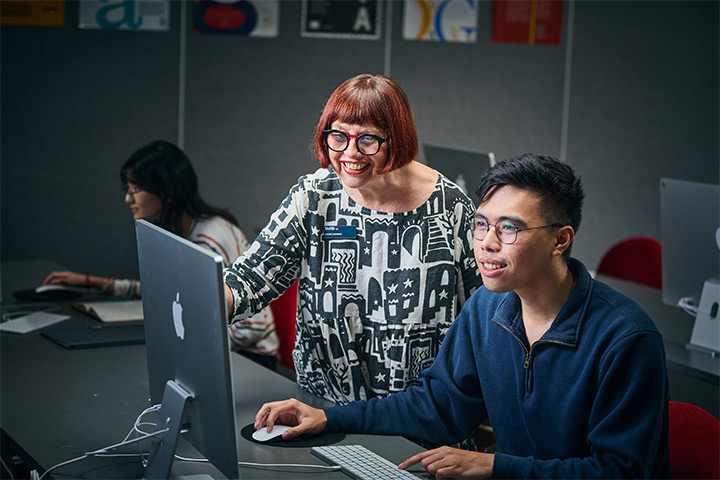 Graphic design student works at a desktop computer while a teacher provides guidance, with design posters on the wall and other students working in the background.