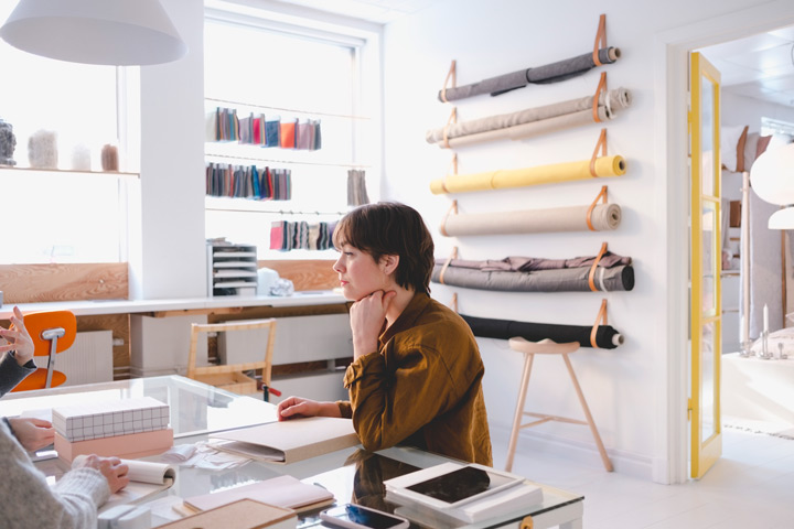 A young woman is sitting in a design studio at a desk. There are reams and swatches of fabric behind her. She is wearing a brown, long-sleeved shirt and looks to be concentrating or listening intently.