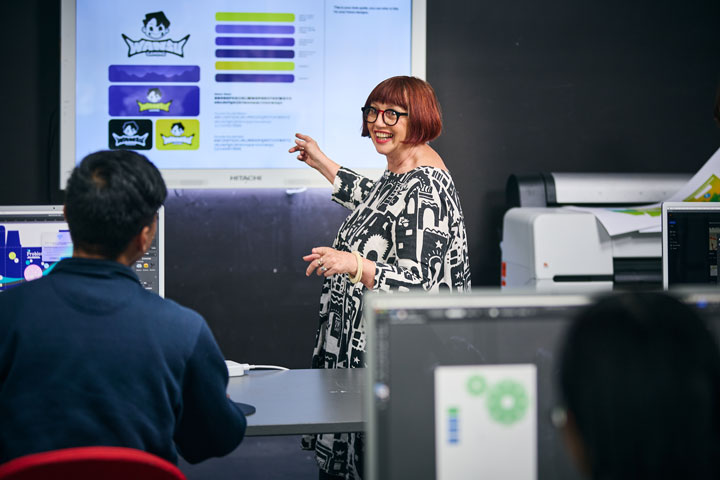 A smiling teacher stands at the front of the class pointing to a smart screen mounted on the wall.