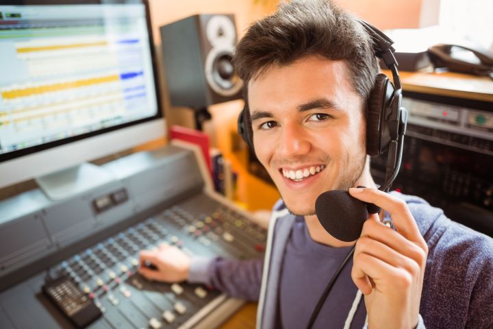 A smiling young man wearing headphones speaks into a microphone at a mixing console in a recording studio.