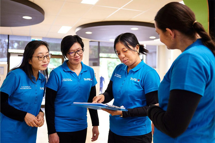 A group of four female childcare workers stand in a workspace. They all wear the TAFE NSW blue t-shirt uniform. One of the workers holds a folder while pointing out some notes to the others. They are relaxed and smiling.