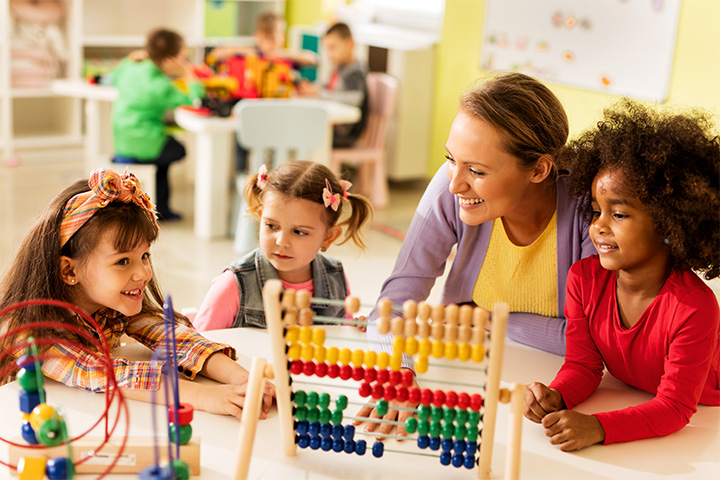 A childcare worker interacts with three young children in a brightly lit classroom. The children are gathered around a colourful wooden abacus, smiling and engaged.
