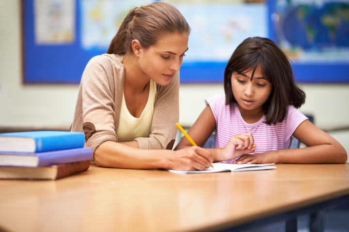 A teacher sitting beside a student at a desk, guiding the student’s hand with a pencil as they write in a notebook. Books are stacked on the desk, and maps and educational posters are visible in the background.