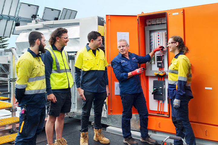 A workshop scene with an electrotechnology teacher guiding four students as they collaborate on a hands-on electrical project.