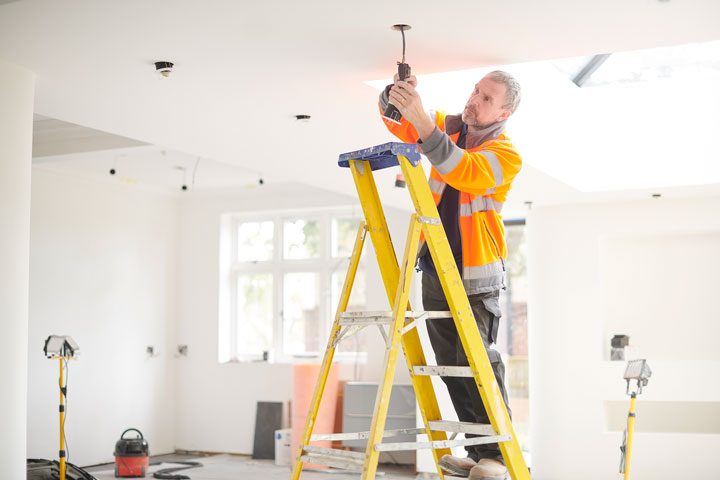 A worker in a high-visibility jacket stands on a yellow ladder installing a light fixture. He is in a bright white room that is being built. Other building equipment is scattered around him including work lights and a vacuum cleaner.