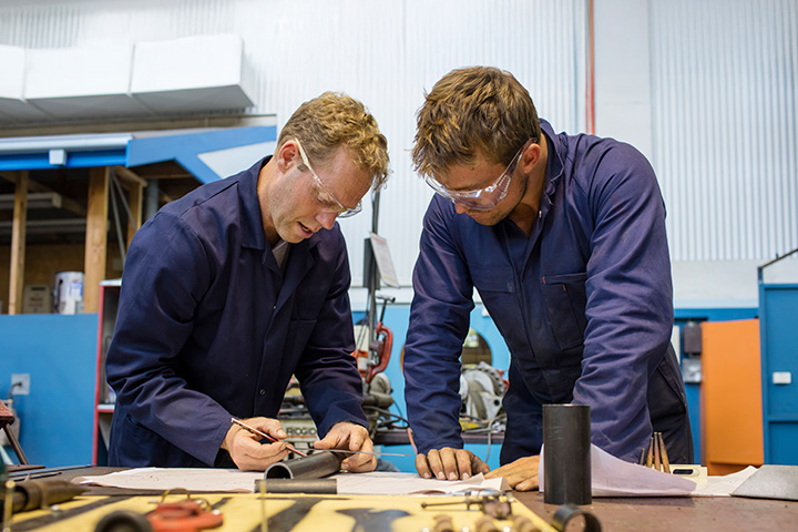 Engineering teacher Craig Walker works alongside a student in a TAFE NSW workshop, reviewing technical drawings and metal components on a workbench surrounded by tools and machinery.