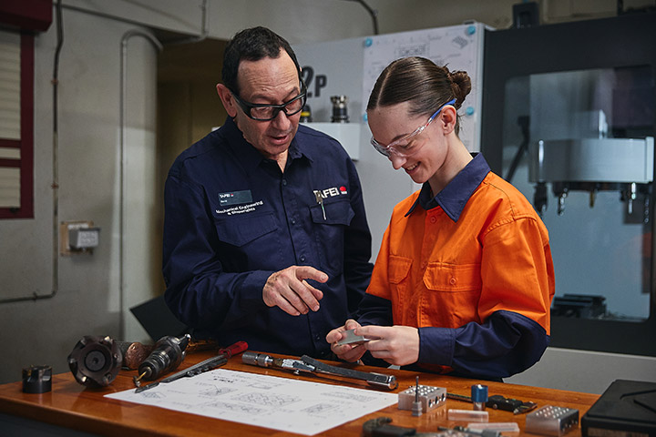 Two people in a workshop, one wearing a navy blue shirt and the other wearing an orange shirt, discussing and examining a mechanical part on a workbench with various tools and technical drawings.