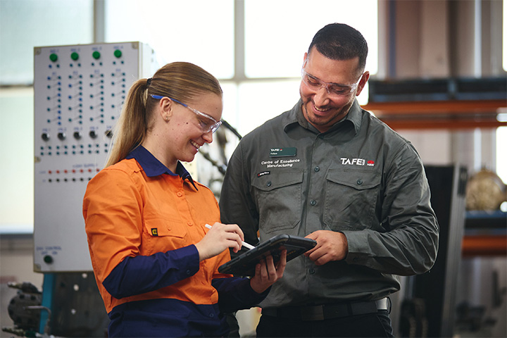 A female manufacturing student in an orange and blue uniform holding a tablet and pointer, shares the tablet screen with her TAFE NSW teacher, who stands beside her.
