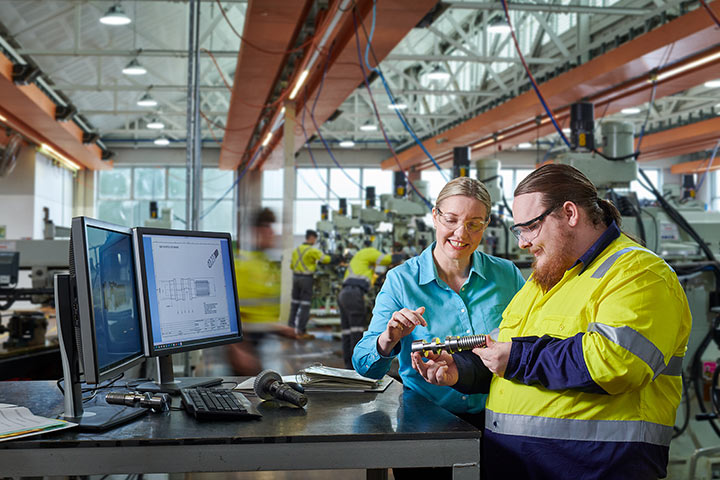 Inside a brightly lit workshop, a male engineering student wearing safety glasses focuses intently on a project, using tools and equipment under the watchful eye of their instructor. In the background, several other students work diligently on their own individual engineering projects.