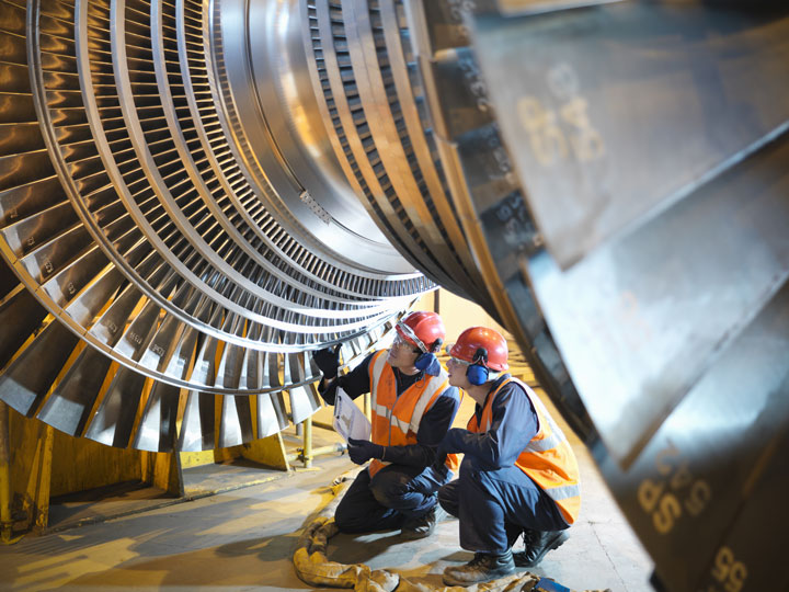 Two engineers crouch to inspect a large turbine, wearing personal protective equipment.