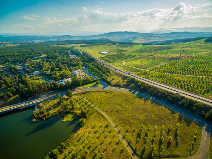 Aerial view of a scenic landscape with roads, a body of water, and green fields lined with rows of trees. In the distance, there are forests, hills, and mountains under a partly cloudy sky.