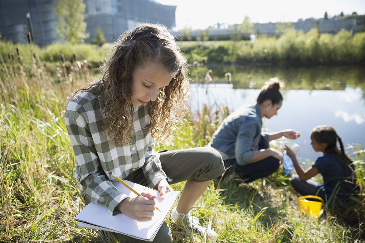 A girl is taking notes while sitting by a body of water. In the background, two people appear to be engaging in an activity related to environmental exploration.