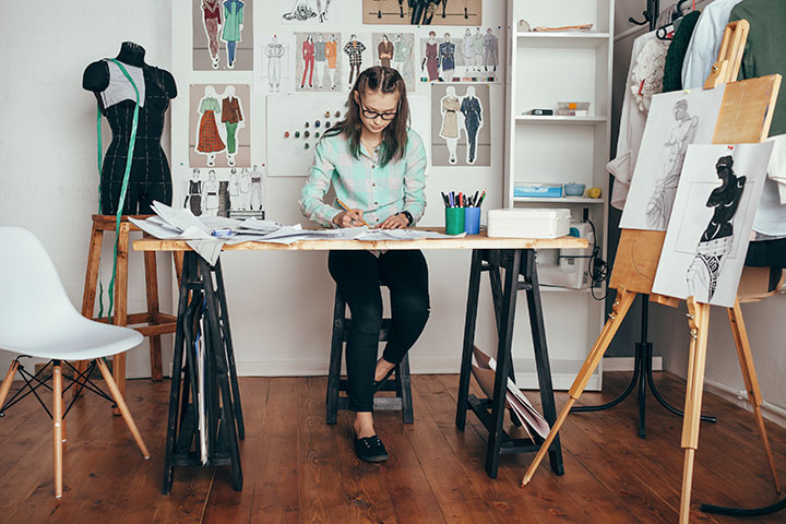 A woman sits at a desk working on some fashion design sketches. There are designs pinned up on the wall behind her, and an art easel is in front to her left. There is a mannequin on her right side with a measuring tape draped over it.