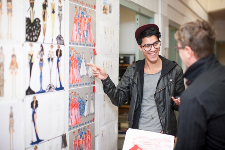 A young man points to fashion design sketches pinned to a wall.