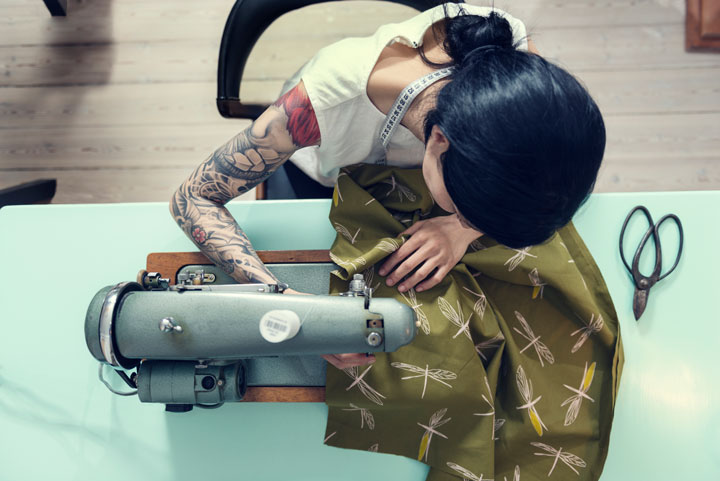 A woman with a tattoo sews green dragonfly-printed fabric at a sewing machine.