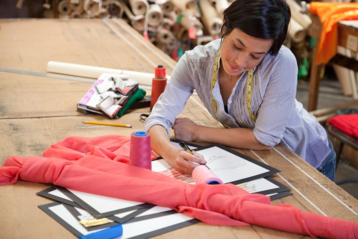 A woman works on fashion design sketches at a wooden table, with materials and design tools around her.