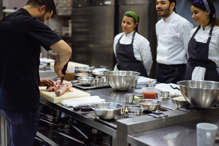 A chef demonstrates how to prepare and cut a piece of meat to three attentive culinary students dressed in aprons and chef uniforms. This is in a commercial kitchen where stainless steel bowls, utensils and ingredients create a busy but organised workspace.