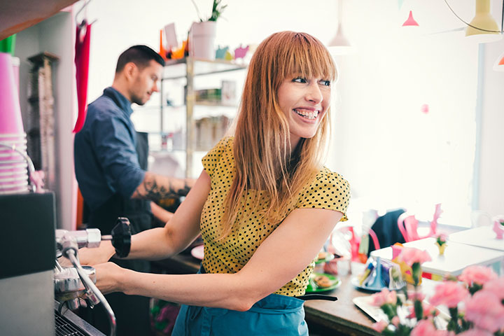 A young lady barista smiling while making coffee at a cafe, turning her head to chat with a customer. In the background, a young man in an apron serves another customer.