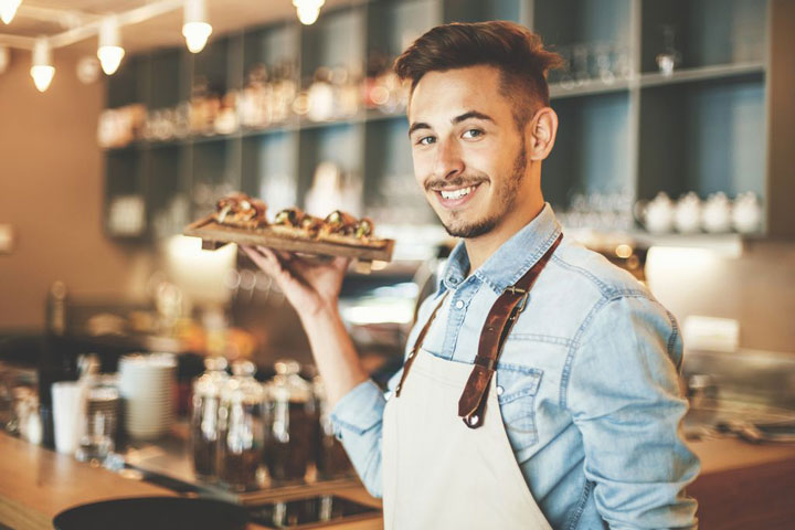 A smiling barista holds a tray of appetizers in a restaurant.