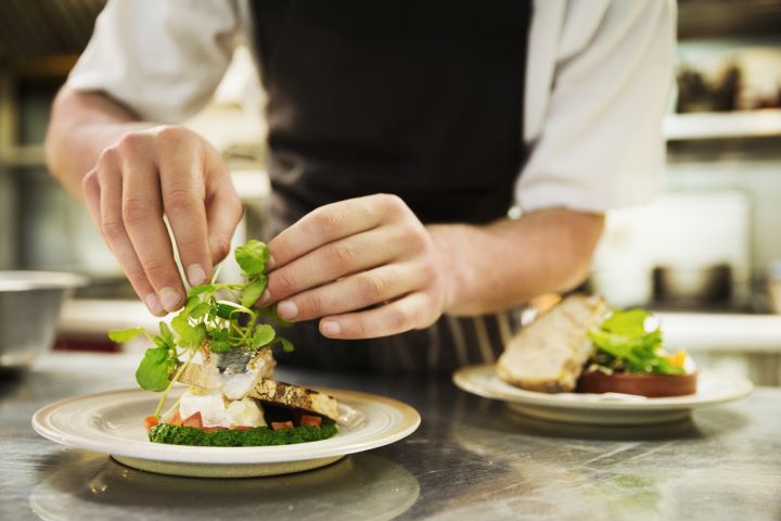 A chef garnishes a plate of grilled fish with herbs. Another plated dish is visible in the background.