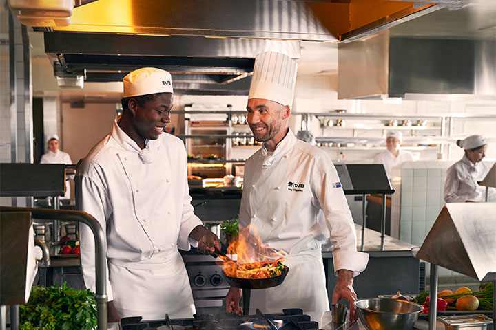 Teacher and student chef collaborating in a bustling commercial kitchen, honing skills and creating culinary masterpieces together.