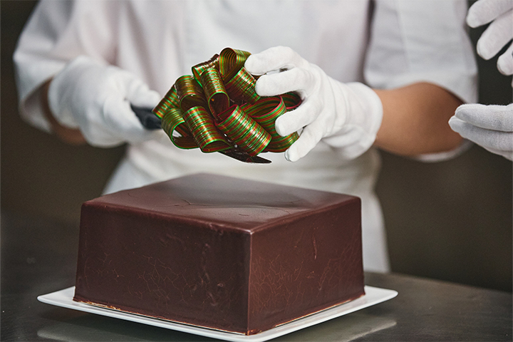 A person in a white chefs uniform and white gloves carefully puts a decorative bow on top of a large square chocolate cake.